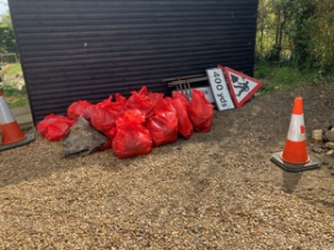 Sacks of rubbish collected piled on gravel at Village Hall car park, Abbotsley, including two traffic cones and roadworks sign.